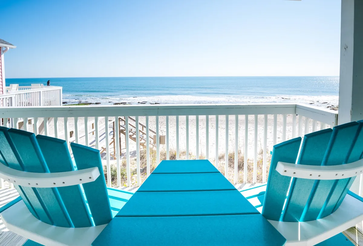 A picturesque view of a white sandy beach framed by turquoise chairs, ocean waves, and open sky. The setting is ideal for sunbathing, reading, building sandcastles, or simply enjoying the tranquil Atlantic shoreline as a family.