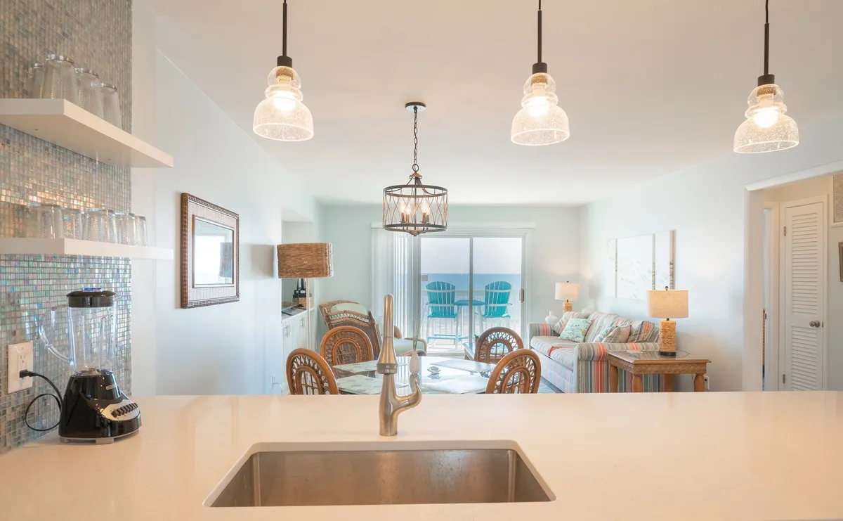 An open-concept living area featuring a kitchen island and hanging light fixtures, with a clear sightline to the living space and a stunning ocean view through large windows. The scene conveys a sense of togetherness, coastal décor, and practical luxury.
