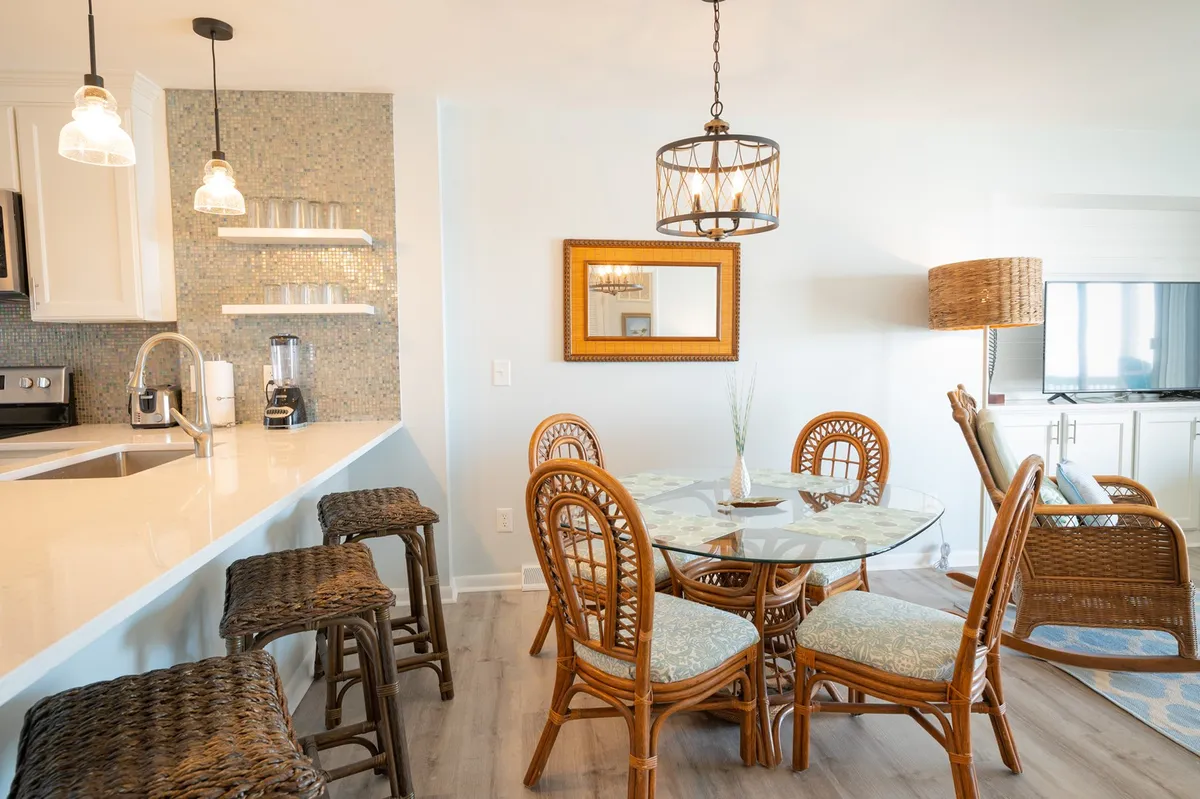 An inviting dining area with a glass dining table surrounded by rattan chairs, elegant pendant lighting, and a mirrored wall decor. The kitchen bar is also visible, connecting the dining and kitchen spaces.