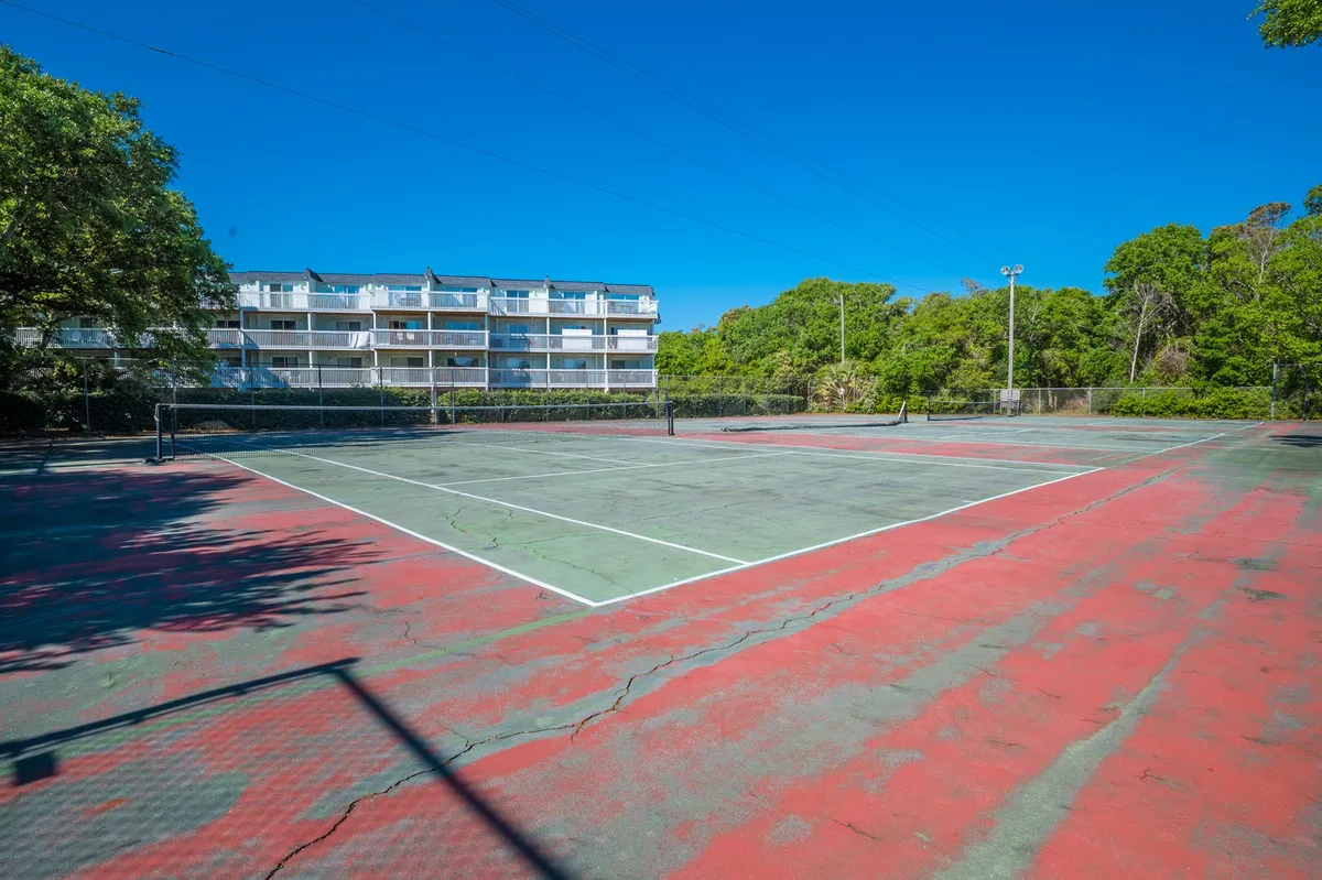 The image features an outdoor tennis court surrounded by greenery, with apartment buildings visible in the background. The well-maintained court is ready for recreational play and friendly competition.