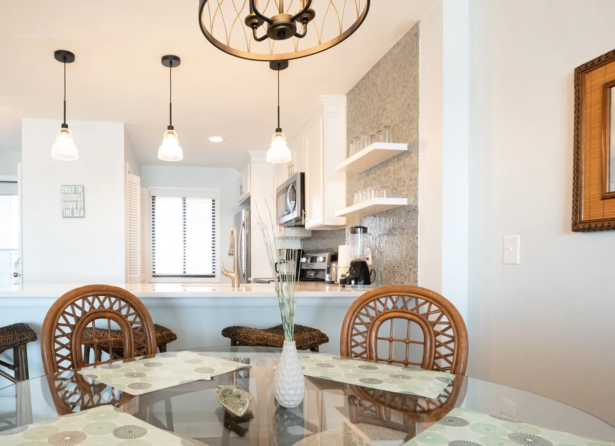 A cozy dining area featuring a glass-top table with rattan chairs, a modern light fixture above, and a decorative vase in the center. The setup encourages relaxed, intimate meals and conversation.