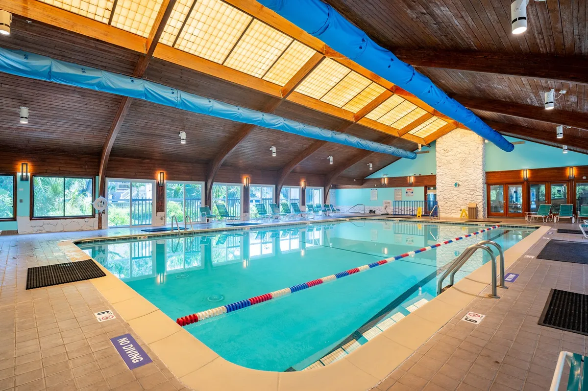 The image shows a spacious indoor pool area with a wooden ceiling and large windows allowing natural light to flood the space. Lounge chairs are set up along the poolside, creating an inviting atmosphere for both relaxation and recreation.