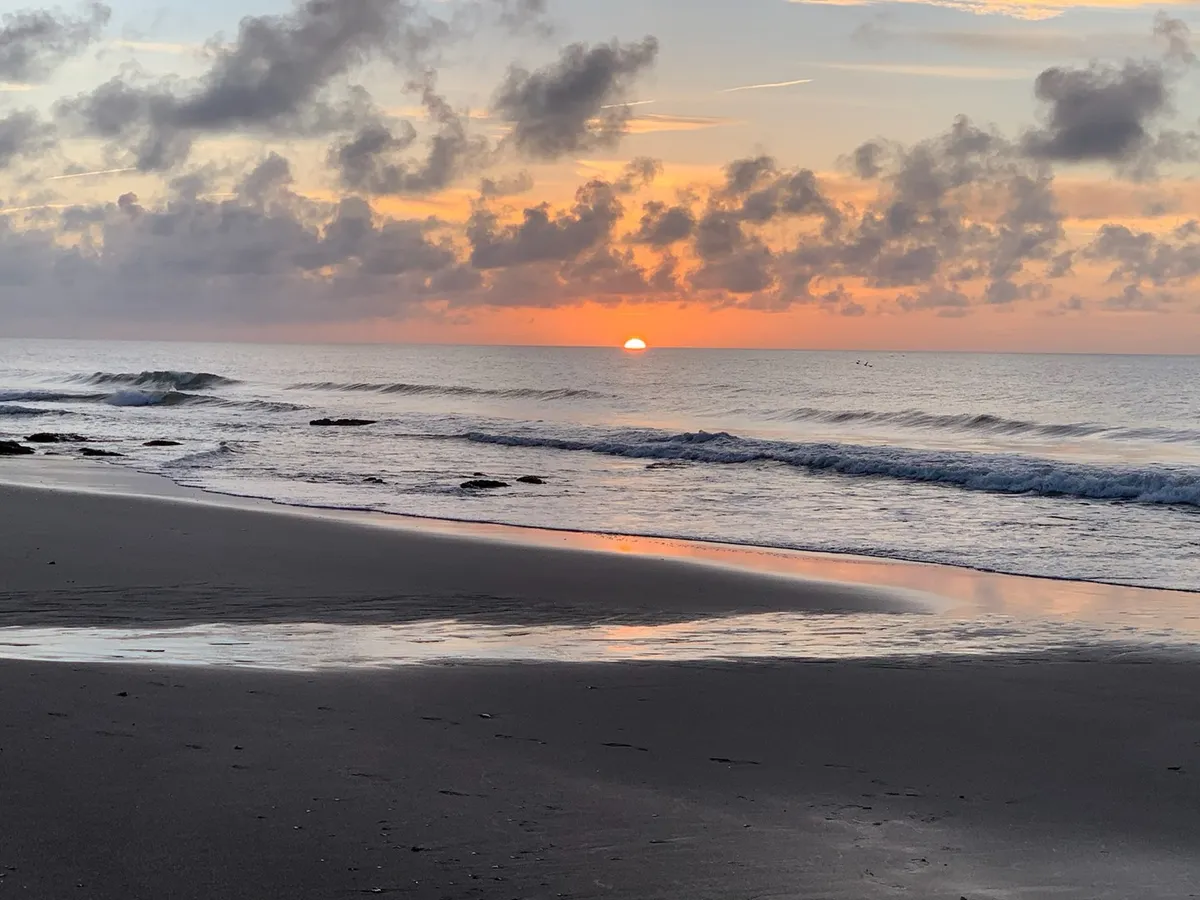 A mesmerizing sunset over the ocean, with gentle waves lapping at wet sand and dramatic clouds painted in warm hues. The scene is calm and reflective, perfectly capturing the magic of dusk on the shore.