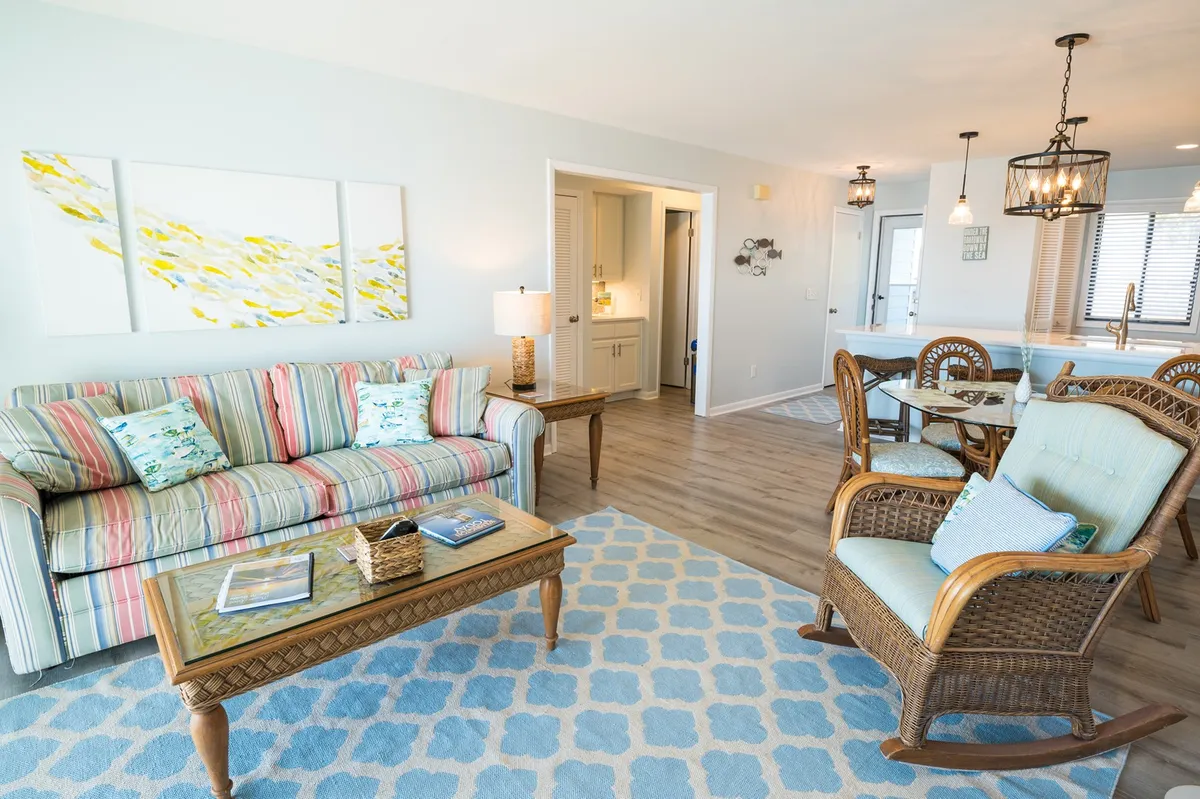 A spacious and comfortable living room featuring a striped sofa (which doubles as a queen sleeper), wooden coffee table, wicker chairs, and beach-themed artwork. The mood is inviting, echoing the comfort and relaxation throughout the property.