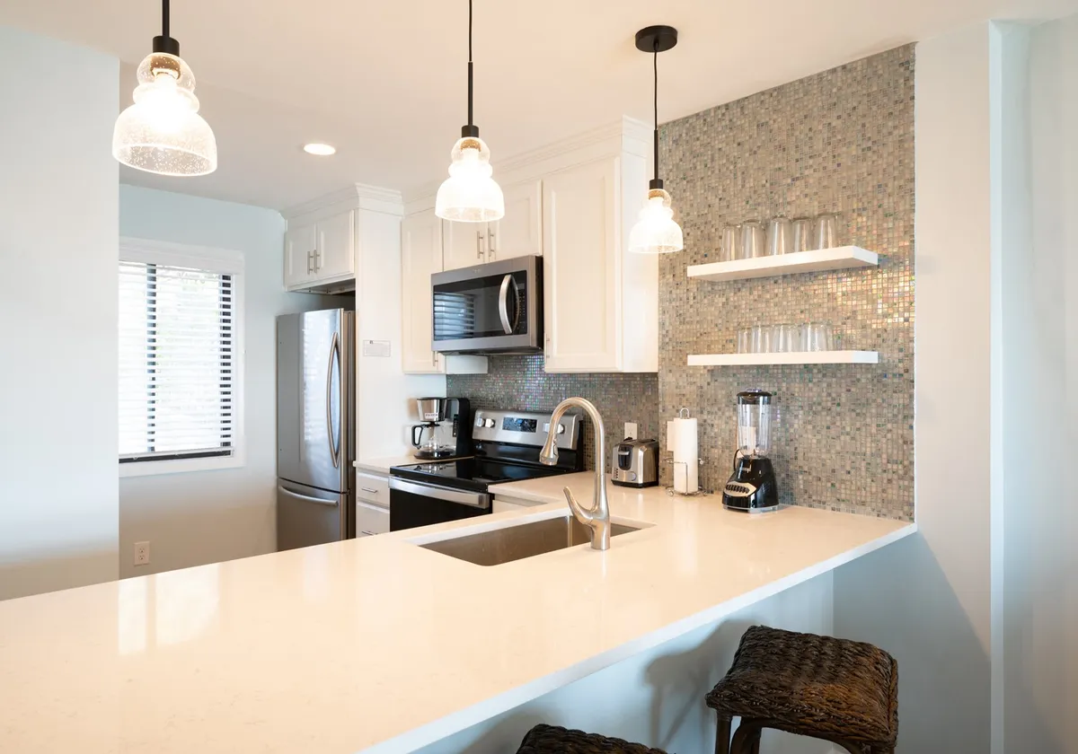 A bright, modern kitchen featuring a spacious island with bar seating for three, pendant lights overhead, sleek backsplash tiles, and upgraded stainless steel appliances. The open layout hints at a welcoming environment ideal for meal prep and socializing.