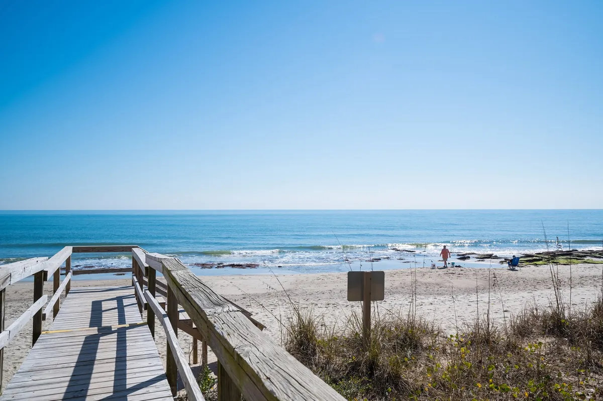 A wide wooden walkway leads out over a sandy beach toward the calm ocean, with a few distant figures strolling along the shoreline. Gentle waves and soft sand create a peaceful, inviting landscape ideal for quiet walks or seaside exploration.
