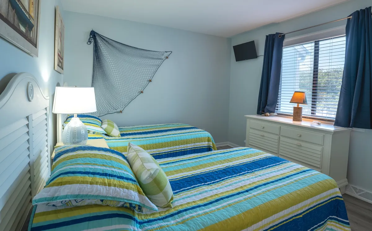 A comfortable guest bedroom with two twin beds adorned with striped quilts, a decorative lamp on the dresser, and sunlight filtering through the curtained window. The atmosphere is cozy and welcoming, suited for children or friends.