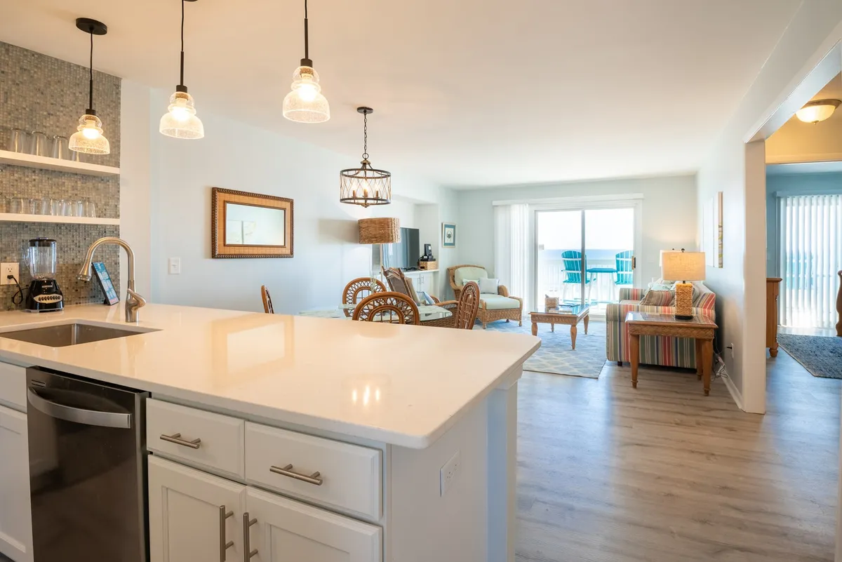 A cozy, welcoming living room that transitions seamlessly to an outdoor deck via large sliding doors. The space includes comfortable seating, a kitchen island with hanging light fixtures, and boasts views perfect for sunset watching or relaxed mornings.