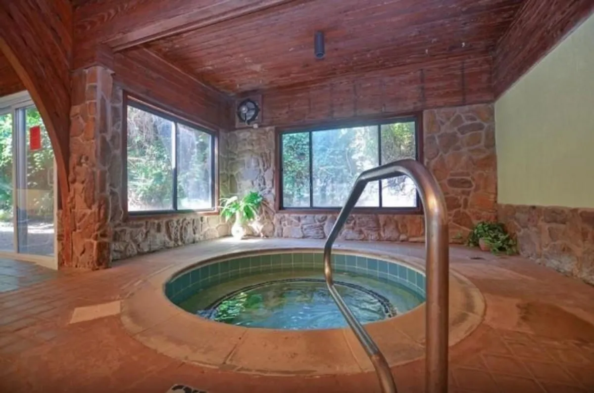 A serene indoor hot tub room with large windows that frame a view of nature. The stone walls, lush plants, and inviting water create a sense of luxury and private relaxation.