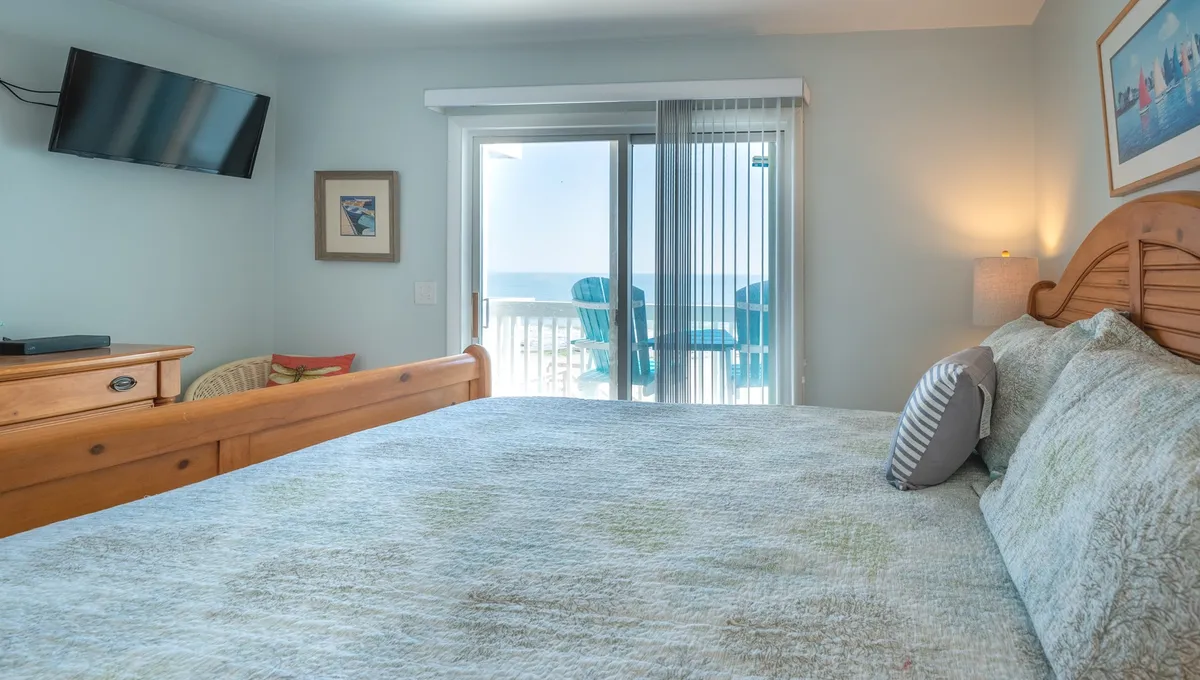 A serene primary bedroom featuring a king bed with plush bedding, soft natural light streaming through sliding glass doors, and artwork adding warmth. The doors open to a private deck with an ocean view, creating a tranquil atmosphere ideal for relaxation.